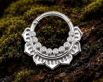 Pode incluir: Um anel circular tipo clicker de tom prateado com uma borda decorativa em forma de leque e sete pequenas pedras transparentes de corte redondo cravadas ao longo da curva interna. O metal é polido e apresenta recortes em forma de pétalas.