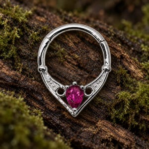 Puede incluir: Un anillo de bisagra en tono plateado con un segmento decorativo en forma de V, adornado con una piedra preciosa central de color rosa intenso en talla pera y peque&ntilde;as piedras claras incrustadas en el marco met&aacute;lico para piercing.
