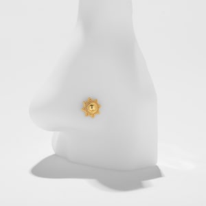 May include: A gold-colored nose stud with a sunburst design. The stud features a central circular element with the letter "T" inside, surrounded by a detailed, beaded border and radiating points. The jewelry is shown on a white mannequin.