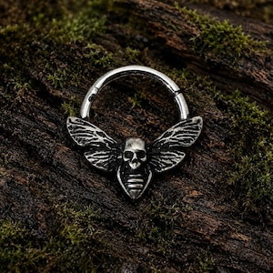 Puede incluir: Un anillo circular articulado de tono plateado con un adorno central en forma de polilla. El diseño incluye una calavera detallada en el centro, alas texturizadas que se extienden hacia los lados y un cuerpo segmentado bajo la calavera.