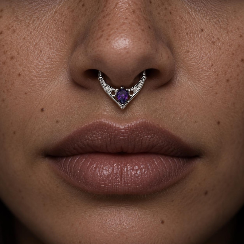 Puede incluir: Un anillo de septum de plata con un dise&ntilde;o en forma de V, detalles met&aacute;licos intrincados y una piedra preciosa morada en forma de pera en el centro. La joya est&aacute; colocada en el tabique nasal y presenta un acabado met&aacute;lico pulido y sim&eacute;trico.