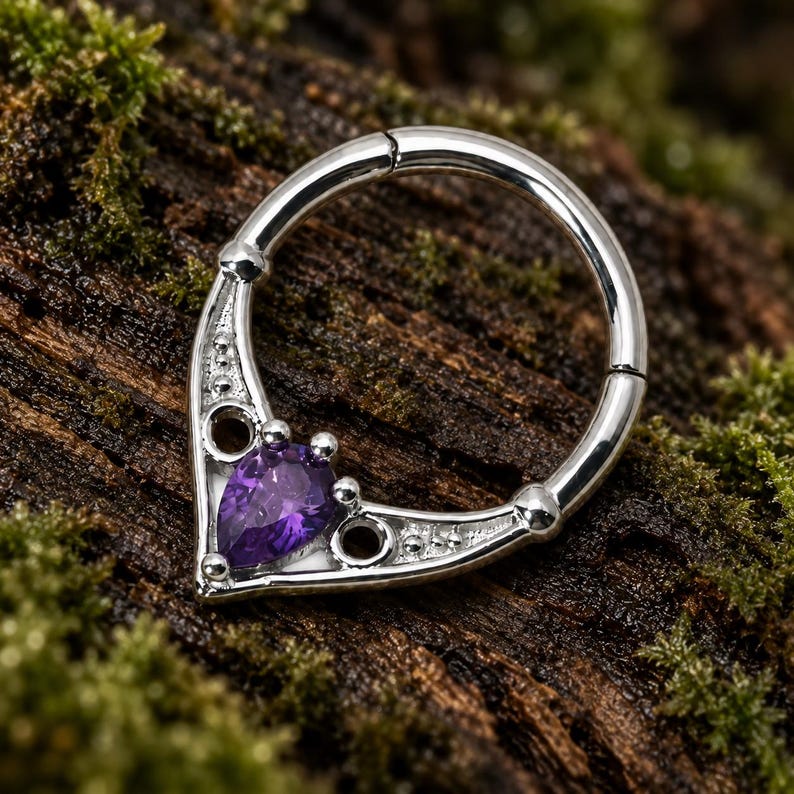 Puede incluir: Un anillo de segmento articulado en tono plateado con un frente decorativo en forma de V. El dise&ntilde;o incluye una piedra preciosa morada central con corte de pera, dos peque&ntilde;os recortes circulares y detalles de cuentas en el marco met&aacute;lico.