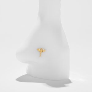 May include: A gold nose stud with a dragon motif, including outstretched wings and a small, clear gemstone. The stud is displayed on a white nose model, highlighting its design and fit. The background is a simple white, emphasizing the jewellery.