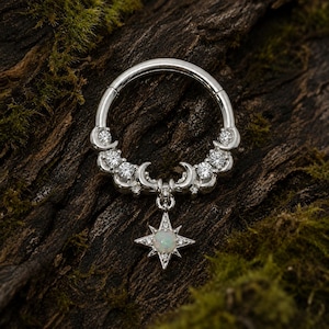 Puede incluir: Un anillo circular de tono plateado con una serie de pequeñas piedras transparentes y detalles de media luna a lo largo de la curva superior. Un dije colgante en forma de estrella cuelga del centro, con un ópalo iridiscente y piedras.
