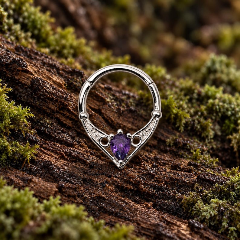 Puede incluir: Un anillo de septum circular de tono plateado con un segmento decorativo en forma de V en la parte inferior. El centro de la forma en V tiene una piedra preciosa morada en forma de pera, flanqueada por dos peque&ntilde;os recortes circulares.