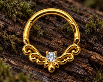 Pode incluir: Um anel circular de cor dourada com um design decorativo de filigrana na metade inferior. O centro do ornamento metálico é cravejado com uma única pedra preciosa transparente de corte redondo que brilha sobre a superfície dourada.