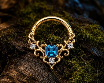 Pode incluir: Um anel de aro dourado com um design de filigrana ornamentado, uma pedra preciosa azul central de corte quadrado e três pequenas pedras de destaque redondas e transparentes. O metal apresenta padrões intrincados que emolduram a pedra azul.