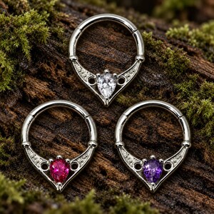 Puede incluir: Tres anillos de segmento articulados en tono plateado con una base decorativa en forma de V. Cada anillo tiene una piedra preciosa en forma de pera en el centro, incluyendo una transparente, una rosa brillante y una morada, con peque&ntilde;os huecos.