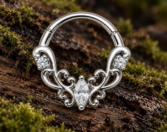 Pode incluir: Um anel circular de tom prateado com um design de metal ornamentado na metade inferior. A peça é adornada com uma pedra preciosa transparente central de corte marquise e duas pedras redondas menores incrustadas nos arabescos.