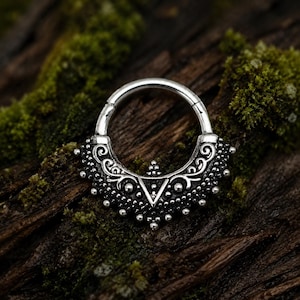 May include: A silver-toned circular hoop ring featuring an ornate, semi-circular decorative attachment. The lower portion is embellished with intricate scrollwork, small raised dots, and a central triangular cutout, creating a detailed metal design.