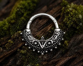 Puede incluir: Un anillo circular de tono plateado con un accesorio decorativo semicircular ornamentado. La parte inferior está adornada con volutas intrincadas, pequeños puntos en relieve y un recorte triangular central en un diseño de metal detallado.