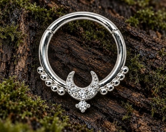 Pode incluir: Um anel circular de tom prateado com um motivo central de meia-lua adornado com pequenas pedras transparentes. O anel inclui um detalhe de pedra pendente na parte inferior e uma série de pequenas contas esféricas ao longo da curva inferior.