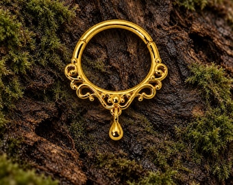 Pode incluir: Um anel circular de cor dourada com um design de filigrana ornamental e simétrico ao longo da metade inferior e um pingente liso em forma de gota suspenso no ponto central da estrutura metálica decorativa.