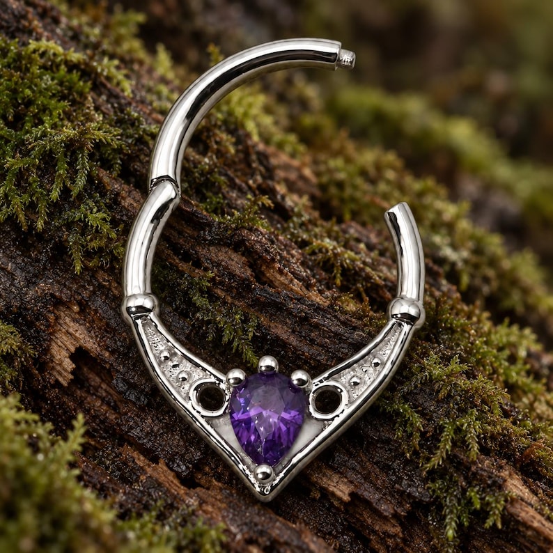Puede incluir: Un anillo de segmento articulado en tono plateado con una base decorativa en forma de V. El centro de la base presenta una piedra preciosa morada en forma de pera, flanqueada por dos peque&ntilde;os recortes circulares y detalles de cuentas met&aacute;licas.