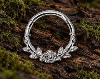 Pode incluir: Um anel circular de tom prateado com um relevo detalhado de uma flor de rosa central, ladeada por motivos simétricos de videiras e folhas ao longo da parte inferior da banda, concebido com um segmento articulado para um fecho seguro.