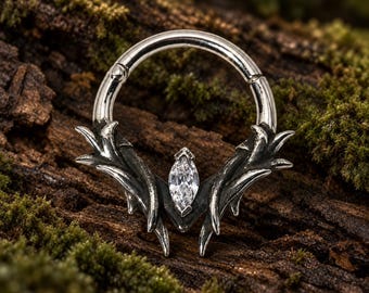 Pode incluir: Um anel circular articulado de tom prateado com uma pedra preciosa transparente central em corte marquise, ladeada por protuberâncias metálicas simétricas em forma de espinho. O metal tem um acabamento polido nas bordas e áreas escurecidas.