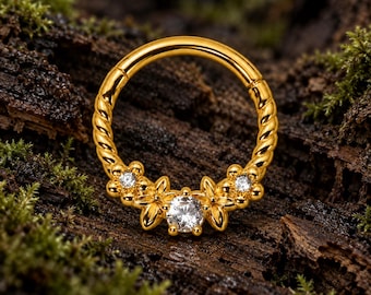 Pode incluir: Um anel circular de tom dourado com um design de banda de metal torcido. A parte inferior é adornada com três engastes em forma de flor, cada um contendo uma pedra preciosa transparente de corte redondo no centro, criando um arranjo floral.