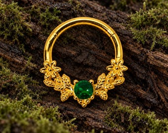 Pode incluir: Um anel circular de tom dourado com um motivo decorativo de flores e folhas na metade inferior. Uma única pedra preciosa redonda de um verde vibrante está cravada centralmente na base, ladeada por flores e folhas metálicas simétricas.