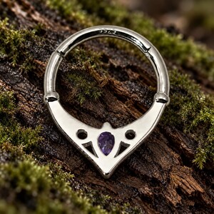 Puede incluir: Un anillo circular articulado de tono plateado con una base decorativa en forma de V con detalles recortados y una piedra preciosa p&uacute;rpura en forma de pera en el centro. La banda de metal tiene grabado el texto S925 en la superficie interior.