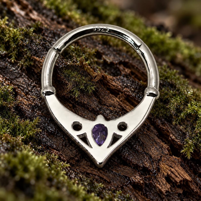 Puede incluir: Un anillo circular articulado de tono plateado con una base decorativa en forma de V con detalles recortados y una piedra preciosa p&uacute;rpura en forma de pera en el centro. La banda de metal tiene grabado el texto S925 en la superficie interior.
