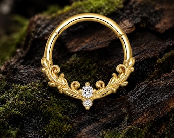 Puede incluir: Un anillo de aro dorado con un diseño de filigrana ornamental y simétrico en la mitad inferior. El trabajo en metal incluye patrones de hojas en espiral y dos piedras transparentes de corte redondo engastadas verticalmente en el centro.
