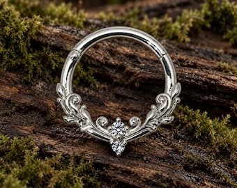 Pode incluir: Um anel circular articulado de tom prateado com um design de filigrana ornamentado na metade inferior. O trabalho em metal inclui padrões de folhas em espiral e duas pequenas pedras redondas transparentes cravadas verticalmente.