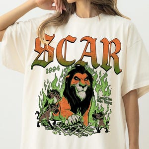 Könnte beinhalten: Ein elfenbeinfarbenes T-Shirt mit einem Grafikdruck eines Löwen mit schwarzer Mähne, drei Hyänen und einem Haufen Knochen, umgeben von grünen Flammen. Der Text SCAR ist in großen orangefarbenen und grünen Buchstaben darüber gedruckt, mit 1994 und Long Live The King.