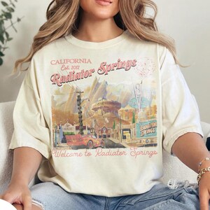May include: Ivory-colored t-shirt featuring a graphic of Radiator Springs, California, with text that reads "Welcome to Radiator Springs." The design includes a red car and various buildings. The shirt is a relaxed fit.