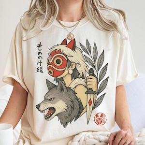May include: Cream-colored t-shirt featuring a graphic of a character with a red mask, a wolf, and a dagger. Japanese text is printed on the left side of the graphic. The shirt is a relaxed fit.