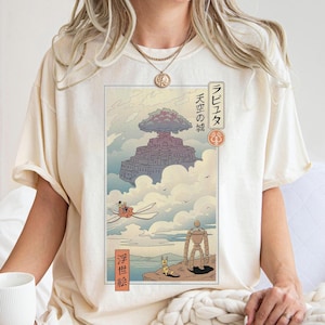 May include: Cream-colored t-shirt featuring a Japanese anime-style illustration. The artwork depicts a floating castle in the clouds, with characters and Japanese text. The shirt has short sleeves and a relaxed fit.