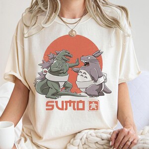 Könnte beinhalten: Cremefarbenes T-Shirt mit einem Aufdruck von Godzilla und Totoro in Sumo-Posen vor einer roten Sonne. Das Wort "SUMO" ist darunter in Orange gedruckt, mit einem japanischen Symbol.