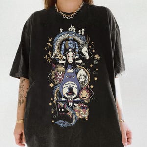 May include: Black t-shirt featuring a colorful graphic of anime characters, including a dragon, Totoro, and other figures. The design is centered on the shirt and includes Japanese text. The shirt has a washed, vintage look.