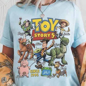 May include: A light blue t-shirt featuring a graphic print of Toy Story characters including Buzz Lightyear, Jessie, Bullseye, Alien, Woody, Rex, Hamm, Slinky, and a green Lilypad with the text Hi!. The design includes the text Toy Story 5 and Since 1995.