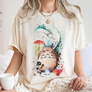 以下が含まれることがあります： 人気アニメのキャラクターの水彩イラストが描かれたクリーム色のTシャツ。赤い傘を持った大きくて茶色い毛むくじゃらの生き物、他のキャラクター、カラフルなアクセントがデザインに含まれています。