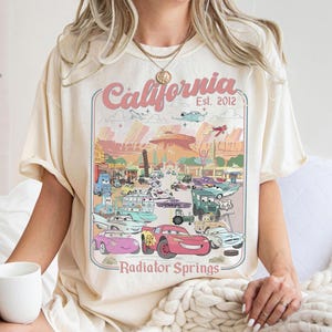 Könnte beinhalten: Cremefarbenes T-Shirt mit einer farbenfrohen Grafik von Zeichentrickautos in Radiator Springs, Kalifornien. Das Design enthält den Text "California Est. 2012" und "Radiator Springs". Das Shirt hat kurze Ärmel und eine lockere Passform.