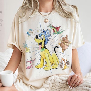 Może przedstawiać: Kremowa koszulka z kreskówkową ilustracją psa Pluto w czapce czarodzieja, otoczonego innymi postaciami z kreskówek. Projekt zawiera niebieski język, zielony kołnierz i promienisty wzór gwiazdy. Koszulka jest noszona przez osobę.