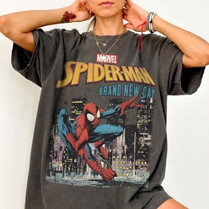 Puede incluir: Camiseta gris oscuro con un gr&aacute;fico de Spider-Man frente a un paisaje urbano. El logotipo de Marvel y las palabras "SPIDER-MAN BRAND NEW DAY" son amarillas y azules. La camiseta es de talla grande.