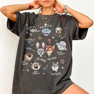 May include: Dark gray t-shirt featuring cartoon character faces from the movie Zootopia. The shirt includes characters like Judy Hopps, Nick Wilde, and Flash. The design is printed on a soft, oversized tee.