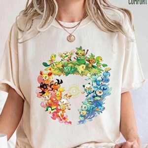 Könnte beinhalten: Ein cremefarbenes Kurzarm-T-Shirt mit einer kreisförmigen Grafik aus verschiedenen bunten Zeichentrickfiguren, die in einem Ring angeordnet sind. Der Text COMFORT COLORS ist in der oberen rechten Ecke aufgedruckt. Rundhalsausschnitt und lockere Passform.