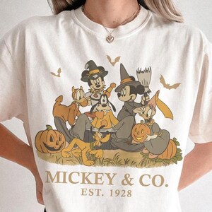 Könnte beinhalten: Ein elfenbeinfarbenes T-Shirt mit einem Grafikdruck von Disney-Figuren in Halloween-Kostümen, darunter Mickey Mouse, Minnie Mouse, Donald Duck und Pluto mit Kürbissen und Fledermäusen. Der Text MICKEY & CO. EST. 1928 steht unter dem Motiv.