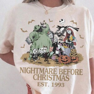 May include: An ivory short-sleeve t-shirt featuring a graphic print of characters from The Nightmare Before Christmas, including Jack Skellington, Sally, and Oogie Boogie. The text reads NIGHTMARE BEFORE CHRISTMAS EST. 1993 with small bats.
