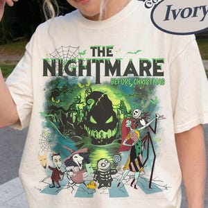 Pode incluir: Uma t-shirt cor de marfim com uma impress&atilde;o gr&aacute;fica de O Estranho Mundo de Jack. O design inclui personagens como Jack Skellington, Sally e Oogie Boogie numa paleta de cores verde e preta com teias de aranha e texto.