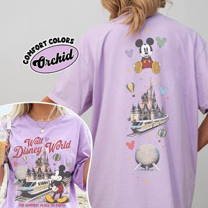 Pu&ograve; includere: Una t-shirt viola chiaro con una stampa grafica di Topolino, il castello di Cenerentola, una monorotaia e Spaceship Earth. Il testo recita Walt Disney World e The Happiest Place on Earth. &Egrave; visibile un'etichetta Comfort Colors Orchid nell'angolo.