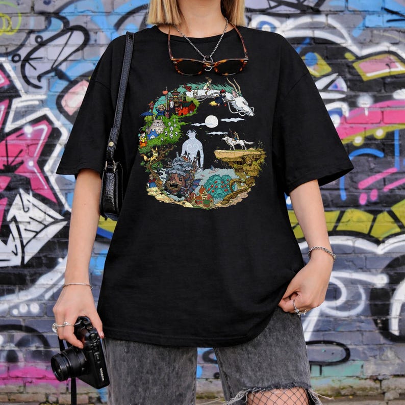 May include: Black t-shirt featuring a colorful graphic design with characters and scenes from an animated film. The design includes a white dragon, a full moon, and various other animated figures. The shirt is worn by a person in front of a graffiti wall.