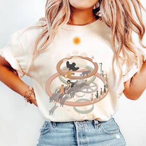 May include: Cream-colored t-shirt featuring a whimsical illustration of cats, gears, and a sun. The design includes a central gear-like structure with cats, a crow, and a person. The shirt has short sleeves and a relaxed fit.