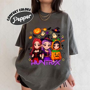 May include: Dark grey t-shirt featuring a Halloween-themed graphic. The design includes three anime-style characters dressed as witches and a pumpkin, with the text "HUNTRX" in purple. The shirt also has the text "COMFORT COLORS Pepper" in a circular design.