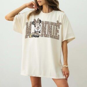 Puede incluir: Camiseta de gran tama&ntilde;o de color crema con la palabra "MINNIE" en una fuente con estampado de leopardo. La letra "O" se reemplaza con un gr&aacute;fico de Minnie Mouse con un vestido y un lazo con estampado de leopardo a juego.