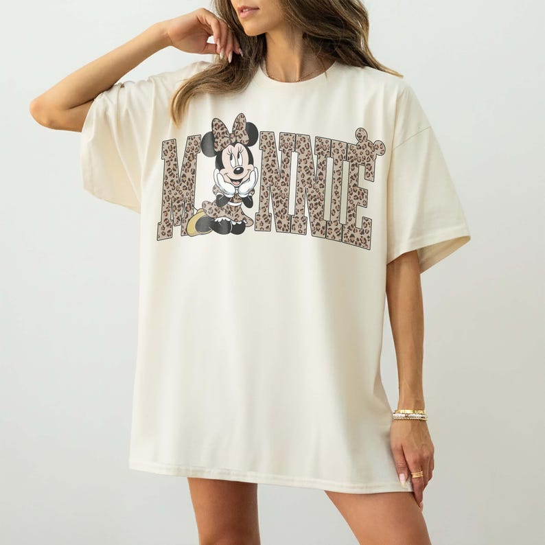 Puede incluir: Camiseta de gran tama&ntilde;o de color crema con la palabra "MINNIE" en una fuente con estampado de leopardo. La letra "O" se reemplaza con un gr&aacute;fico de Minnie Mouse con un vestido y un lazo con estampado de leopardo a juego.