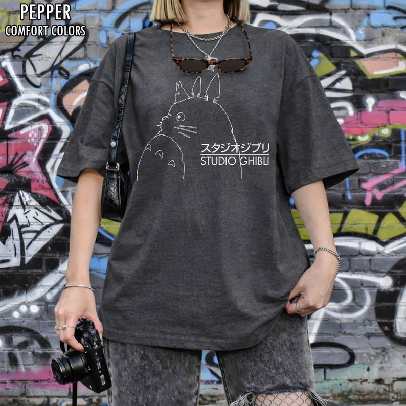 May include: Dark gray t-shirt featuring a white outline of a Studio Ghibli character with Japanese text. The shirt has short sleeves and a relaxed fit. The words "PEPPER" and "COMFORT COLORS" are printed at the top left.