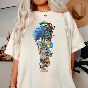 May include: Cream-colored t-shirt featuring a colorful graphic design. The graphic is a collage of animated characters and scenes, with a blue and green element on the left side. The shirt has short sleeves and a crew neck.